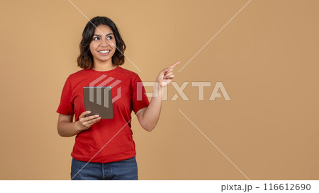 A young middle eastern woman, wearing a red t-shirt and blue jeans, is holding a tablet and pointing her right hand to copy space while smiling. She is standing against a light brown background. 116612690