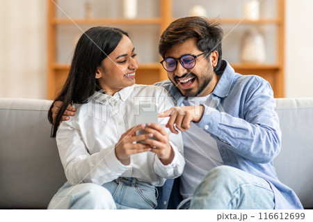 Indian man and woman are seated on a couch, engrossed in their cell phone. The couple is focused on the screen, scrolling and interacting with the device. 116612699