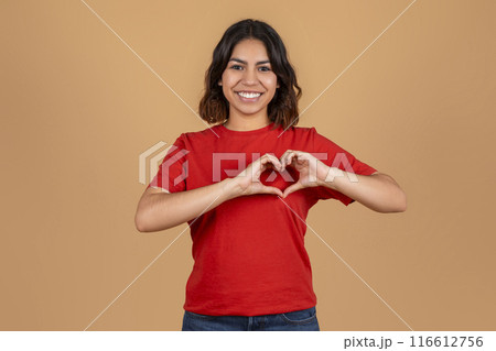 A young middle eastern woman with brown hair and a warm smile, is wearing a red t-shirt and making a heart shape with her hands in front of a light brown background. A young middle eastern woman with brown hair and a warm smile, is wearing a red t-shirt and making a heart shape with her hands in front of a light brown background. 116612756