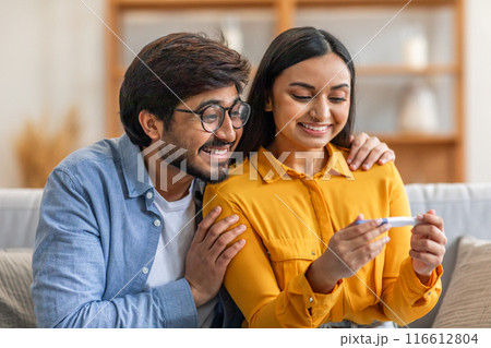 A joyful Indian couple is sitting together on their couch, delightedly examining a positive pregnancy test result in their living room during the daytime. 116612804