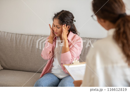 A woman sits on a couch, her head in her hands, appearing anxious during a therapy session. The therapist sits beside her, looking concerned. A woman sits on a couch, her head in her hands, appearing anxious during a therapy session. The therapist sits beside her, looking concerned. 116612819