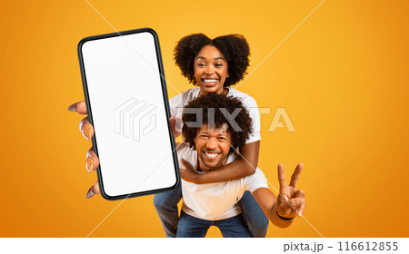 A happy black couple is holding a smartphone with a blank screen in front of a bright yellow background. The woman is giving a peace sign with her right hand and is smiling brightly. A happy black couple is holding a smartphone with a blank screen in front of a bright yellow background. The woman is giving a peace sign with her right hand and is smiling brightly. 116612855