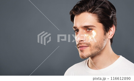 Young man with golden patches under eyes from wrinkles and dark circles, grey studio background 116612890