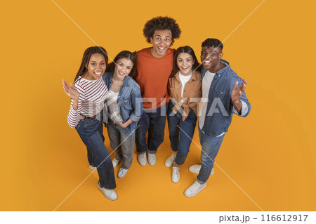 A group of five friends, all dressed in casual attire, pose together on a bright yellow background. They look happy and excited, with their arms around each other, view above 116612917