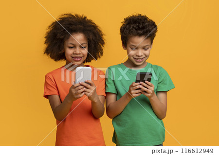 Two African American children, a girl and a boy, are standing in front of a yellow background, smiling while looking down at their smartphones. Two African American children, a girl and a boy, are standing in front of a yellow background, smiling while looking down at their smartphones. 116612949