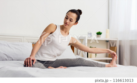 A woman wearing a white tank top and grey leggings stretches her legs while sitting on a bed in a bright room. 116613013