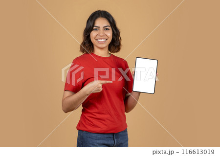 A young middle eastern woman with a bright smile is holding a tablet with a blank white screen. She is wearing a red t-shirt and blue jeans. The background is a light brown color. A young middle eastern woman with a bright smile is holding a tablet with a blank white screen. She is wearing a red t-shirt and blue jeans. The background is a light brown color. 116613019