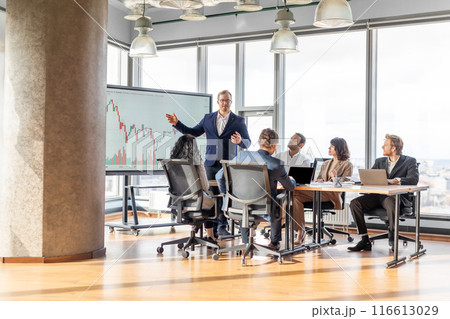 A businessman in a suit stands in front of a whiteboard displaying a financial chart, presenting to a small group of colleagues seated around a table in a modern office with large windows. A businessman in a suit stands in front of a whiteboard displaying a financial chart, presenting to a small group of colleagues seated around a table in a modern office with large windows. 116613029