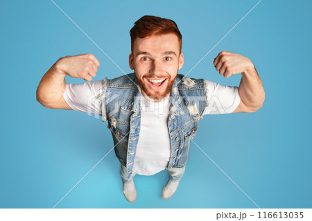 Handsome young redhead bearded man showing his power on blue background, view from above, studio shot Handsome young redhead bearded man showing his power on blue background, view from above, studio shot 116613035