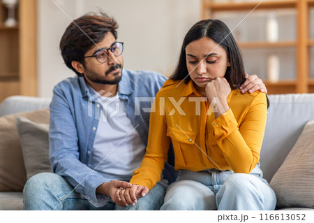 Indian man with glasses and a blue shirt sits on a couch beside a woman in a yellow top, comforting her as she appears upset. He holds her hand and gently rests his other hand on her shoulder 116613052