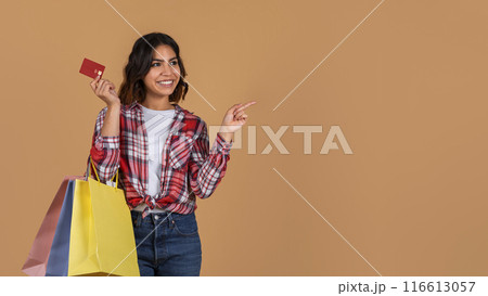 A young arab woman with short, wavy brown hair smiles at the camera while holding a credit card and several shopping bags, pointing at copy space A young arab woman with short, wavy brown hair smiles at the camera while holding a credit card and several shopping bags, pointing at copy space 116613057