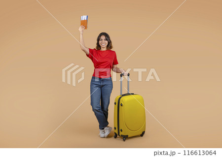 A young middle eastern woman, stands in front of a peach-colored background holding a passport and airplane tickets in one hand and a yellow suitcase in the other. 116613064