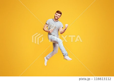 Celebrating Win. Lucky Young Man Jumping On Orange Studio Background With Money In His Hand, Gesturing 116613112