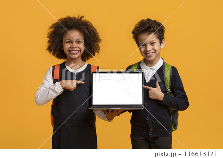 Two African American students, one boy and one girl, stand in front of a bright yellow background. They both wear school uniforms and hold a laptop with a blank screen in front of them 116613121