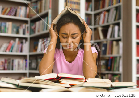 Young black girl student under mental pressure, reading book preparing for examination in library at university, free space 116613129