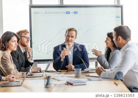 A group of business professionals are seated around a conference table, engaged in a discussion. They are gathered in a modern office with a large whiteboard behind them 116613303