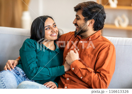 A smiling Indian couple sits on a comfortable sofa in a cozy living room, sharing a heartfelt moment. They are engaging in a loving conversation, enjoying each others company during the day. 116613326