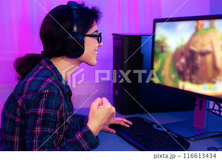 A young woman is pictured playing a video game on a desktop computer. She is wearing a blue and red plaid shirt and headphones and is excitedly pumping her fist in the air. 116613434