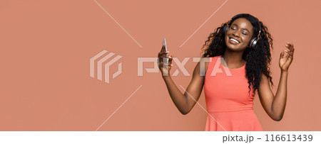 A young black woman with long curly hair is dancing and listening to music on headphones while holding a smartphone in her hand. She is smiling and enjoying the music. 116613439
