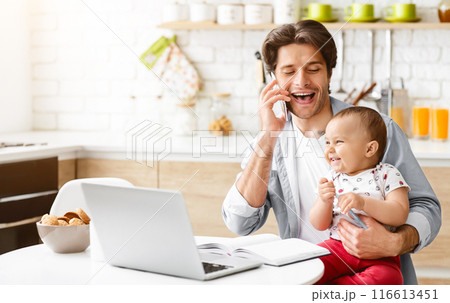 A father is sitting at a kitchen table with his laptop in front of him. He is talking on the phone and smiling while holding his baby. The baby is also smiling and looking at copy space 116613451