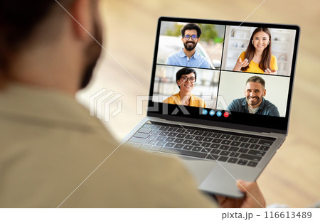 A man sitting at a table is using a laptop, which displays a group of people on the screen. He appears engaged in a virtual meeting or video conference with his colleagues. 116613489