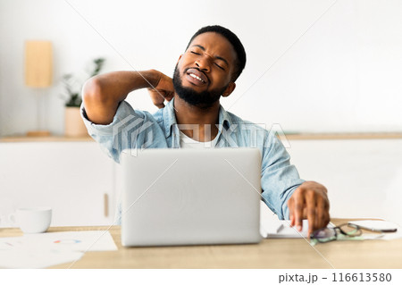 A man sits at a desk with a laptop open in front of him, stretching his neck and looking tired. A man sits at a desk with a laptop open in front of him, stretching his neck and looking tired. 116613580