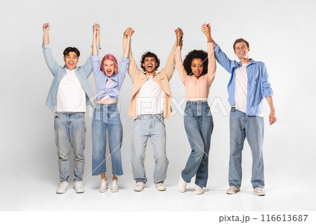 A group of five diverse young adults stand in a studio setting with their arms raised, holding hands and smiling joyfully. They are dressed casually in jeans and shirts. 116613687