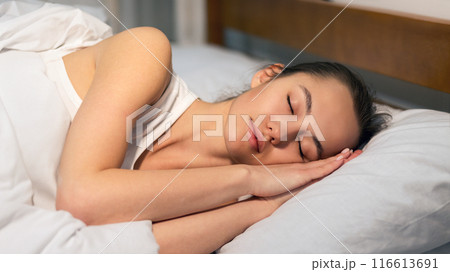 A young woman is sleeping soundly in bed. She is lying on her side with her head resting on a pillow. A young woman is sleeping soundly in bed. She is lying on her side with her head resting on a pillow. 116613691