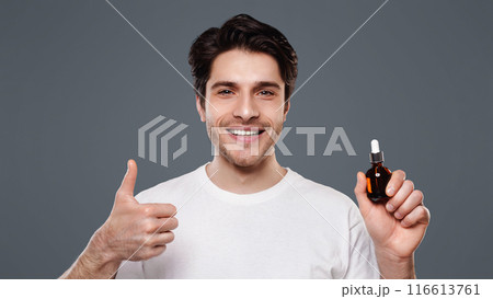 Masculine beauty. Handsome man approving face serum, showing thumb up gesture with small bottle, grey background 116613761