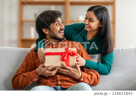 Indian man is seated on a couch, holding a gift box, while a woman sits beside him. The man and woman are indoors, with neutral decor in the background. 116613843