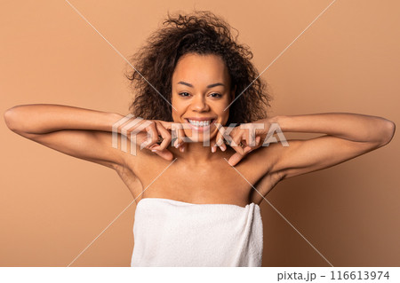 A young African American woman with curly hair is wrapped in a white towel, smiling and looking directly at the camera. She is standing in front of a light brown background. A young African American woman with curly hair is wrapped in a white towel, smiling and looking directly at the camera. She is standing in front of a light brown background. 116613974