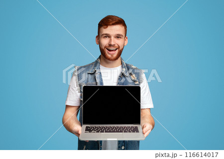 Happy millennial man showing to camera laptop with blank black screen for mockup, isolated on blue studio background 116614017