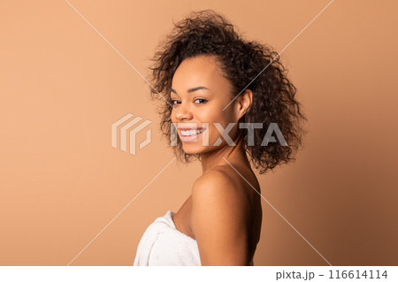 This image shows a young black woman with curly dark brown hair, smiling as she looks over her shoulder at the camera. She is wrapped in a white towel and is standing against a tan background. This image shows a young black woman with curly dark brown hair, smiling as she looks over her shoulder at the camera. She is wrapped in a white towel and is standing against a tan background. 116614114