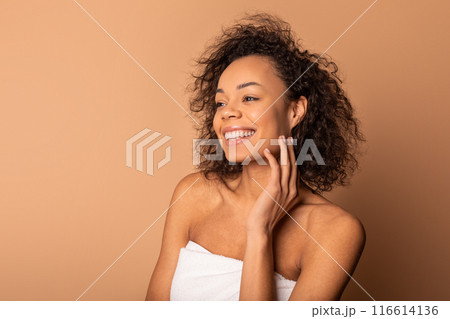 African American smiling woman with curly brown hair is wearing a white towel and looking to the side. She is touching her cheek with her hand. She is standing against a plain brown background. African American smiling woman with curly brown hair is wearing a white towel and looking to the side. She is touching her cheek with her hand. She is standing against a plain brown background. 116614136