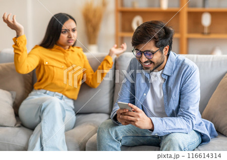 Indian man wearing glasses and a blue shirt is smiling and looking at his phone. Next to him, a woman in a yellow shirt is sitting with raised hands, displaying frustration Indian man wearing glasses and a blue shirt is smiling and looking at his phone. Next to him, a woman in a yellow shirt is sitting with raised hands, displaying frustration 116614314