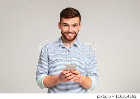 Young bearded man using his phone and smiling on studio background, websurfing or scrolling, copy space 116614362