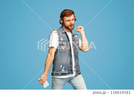 Redhaired stylish millennial man enjoying beloved music and dancing on blue studio background, copy space 116614381
