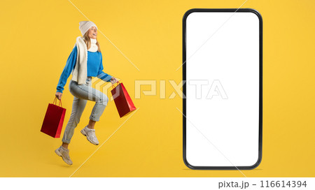 A joyful young woman in casual winter attire holds red shopping bags, leaping near a large smartphone screen with a blank display against a bright yellow backdrop. 116614394