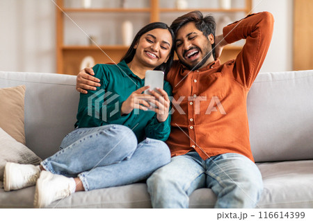 Indian man and woman are seated on a couch, both engrossed in a cell phone. They appear focused on the screen, possibly sharing content or engaging in a conversation related to the device. Indian man and woman are seated on a couch, both engrossed in a cell phone. They appear focused on the screen, possibly sharing content or engaging in a conversation related to the device. 116614399