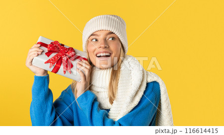 A woman wearing a blue sweater is holding a wrapped present in her hands. She appears to be standing, with a neutral expression on her face. 116614415