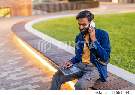Indian man in a blue shirt and yellow t-shirt is sitting on a wooden bench in a park. He is engaged in conversation on his mobile phone while simultaneously working on his laptop Indian man in a blue shirt and yellow t-shirt is sitting on a wooden bench in a park. He is engaged in conversation on his mobile phone while simultaneously working on his laptop 116614446