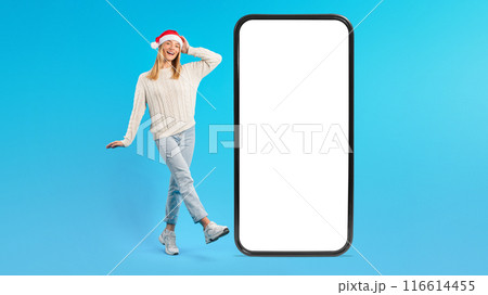 A cheerful young woman wearing a Santa hat and festive attire stands next to a large smartphone with white blank screen, posing playfully against a bright blue background. A cheerful young woman wearing a Santa hat and festive attire stands next to a large smartphone with white blank screen, posing playfully against a bright blue background. 116614455
