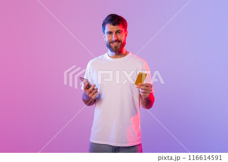 A man is holding a card in one hand and a cell phone in the other. He appears to be focusing on the phone screen while holding the card. The background is neutral and nondescript. A man is holding a card in one hand and a cell phone in the other. He appears to be focusing on the phone screen while holding the card. The background is neutral and nondescript. 116614591