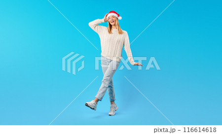 A young woman is standing on one leg, wearing a cozy sweater, jeans, and sneakers, all while donning a Santa hat. She has a joyful expression and her hand on her head A young woman is standing on one leg, wearing a cozy sweater, jeans, and sneakers, all while donning a Santa hat. She has a joyful expression and her hand on her head 116614618