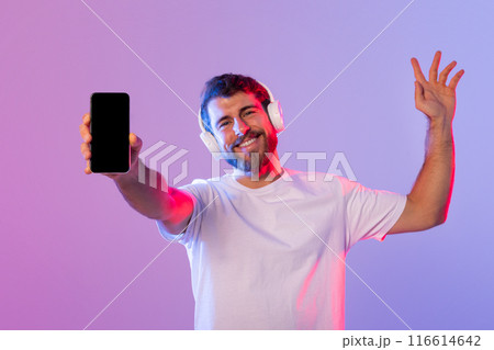 A man wearing headphones holds up a cell phone, with a focused expression on his face. He appears to be listening to music or talking on the phone. A man wearing headphones holds up a cell phone, with a focused expression on his face. He appears to be listening to music or talking on the phone. 116614642