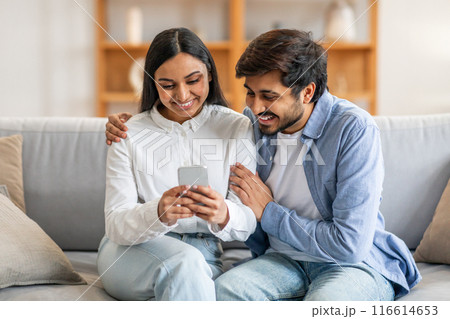 Indian man and woman are seated on a couch, both looking intently at a cell phone screen. They appear engaged in whatever is displayed on the device, with focused expressions on their faces. 116614653