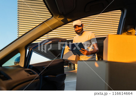 Indian delivery worker wearing a cap and t-shirt is unloading cardboard boxes, from a car at a modern building while holding a clipboard during sunset. Indian delivery worker wearing a cap and t-shirt is unloading cardboard boxes, from a car at a modern building while holding a clipboard during sunset. 116614664