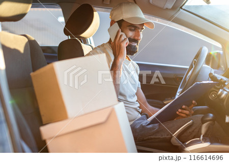 Indian delivery driver is sitting in his car, talking on the phone, and holding a clipboard. Various packages are visible on the passenger seat beside him 116614696