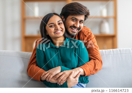 A happy Indian couple is sitting on a couch, with the man embracing the woman from behind. Both are smiling warmly, creating a heartwarming and affectionate atmosphere in a cozy living room. 116614723