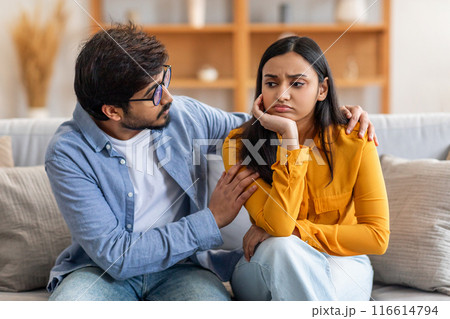 Indian young man and woman sit closely on a comfortable living room couch. The man is gently comforting the woman, who appears to be upset, showing support and understanding. 116614794
